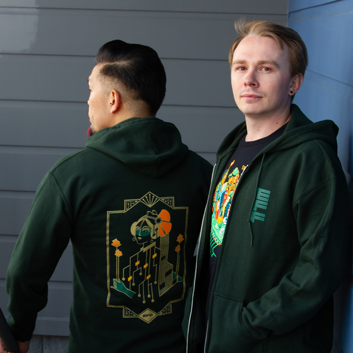 Two people wearing green hooded EFF sweatshirts with a gold "Motherboard" design on the back.