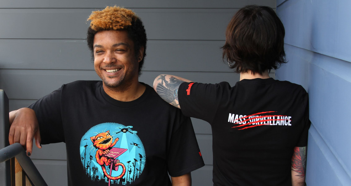 Two people wear EFF Claw Back member t-shirts. The front shows a cat swatting at spy cameras and the back says “Mass Surveillance” with red claw marks through it