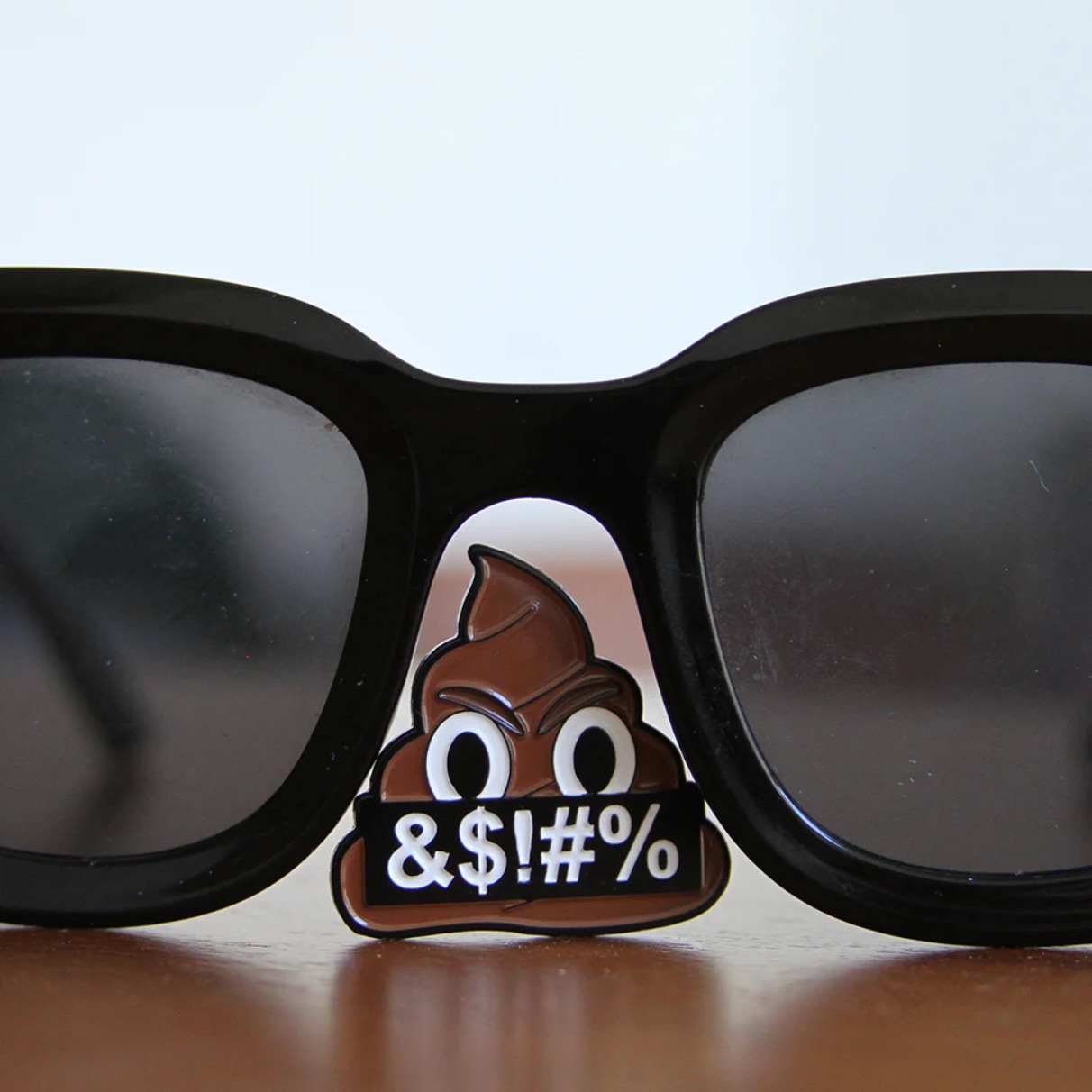 An enamel pin clipped to the nose bridge of black-framed sunglasses resting on a wooden surface. The pin depicts a scowling poop emoji holding a black banner reading "&$!#%".