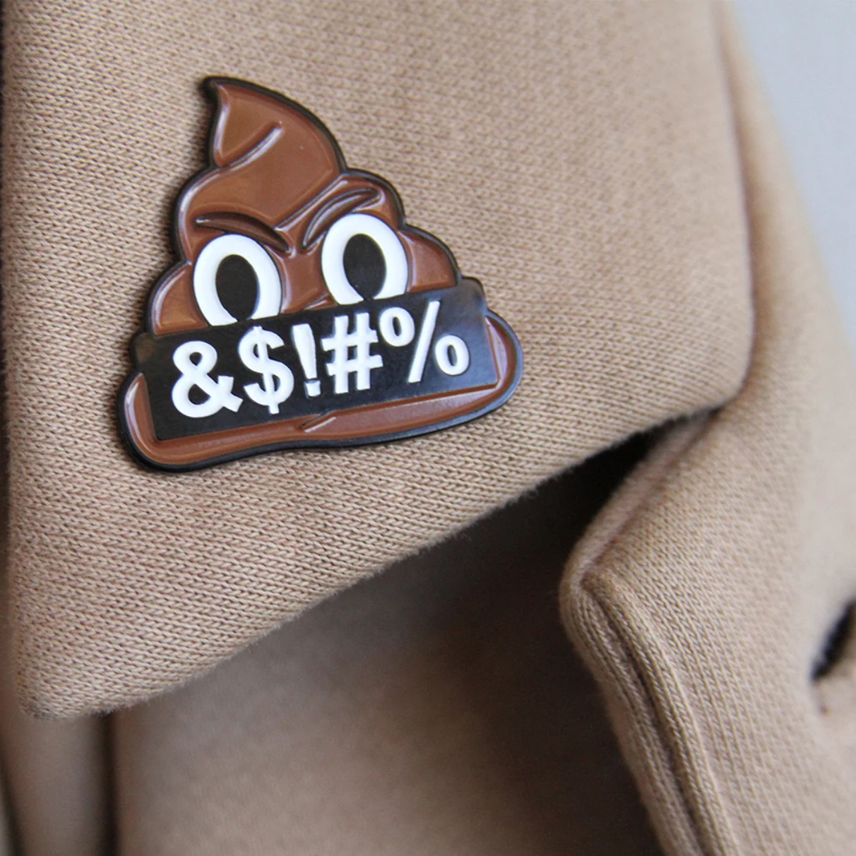 A close-up of an enamel pin on the lapel of a tan jacket. The pin depicts an angry poop emoji holding a black banner reading "&$!#%".