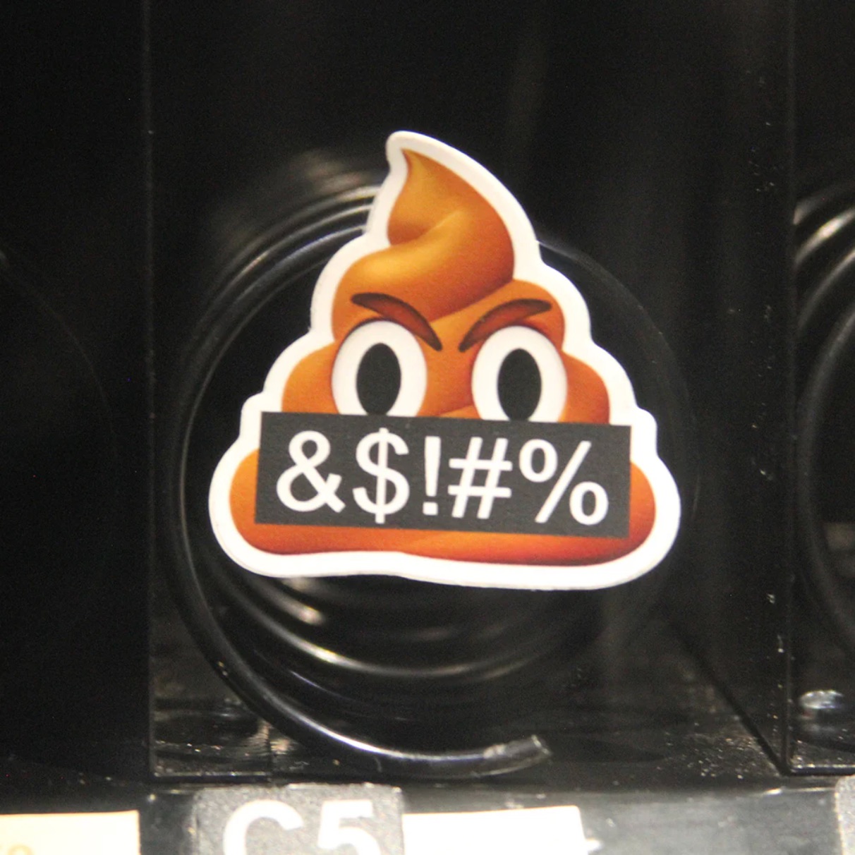 An angry poop emoji sticker affixed to the coiled spring mechanism inside a vending machine. The sticker depicts a scowling poop emoji holding a black sign reading "&$!#%".