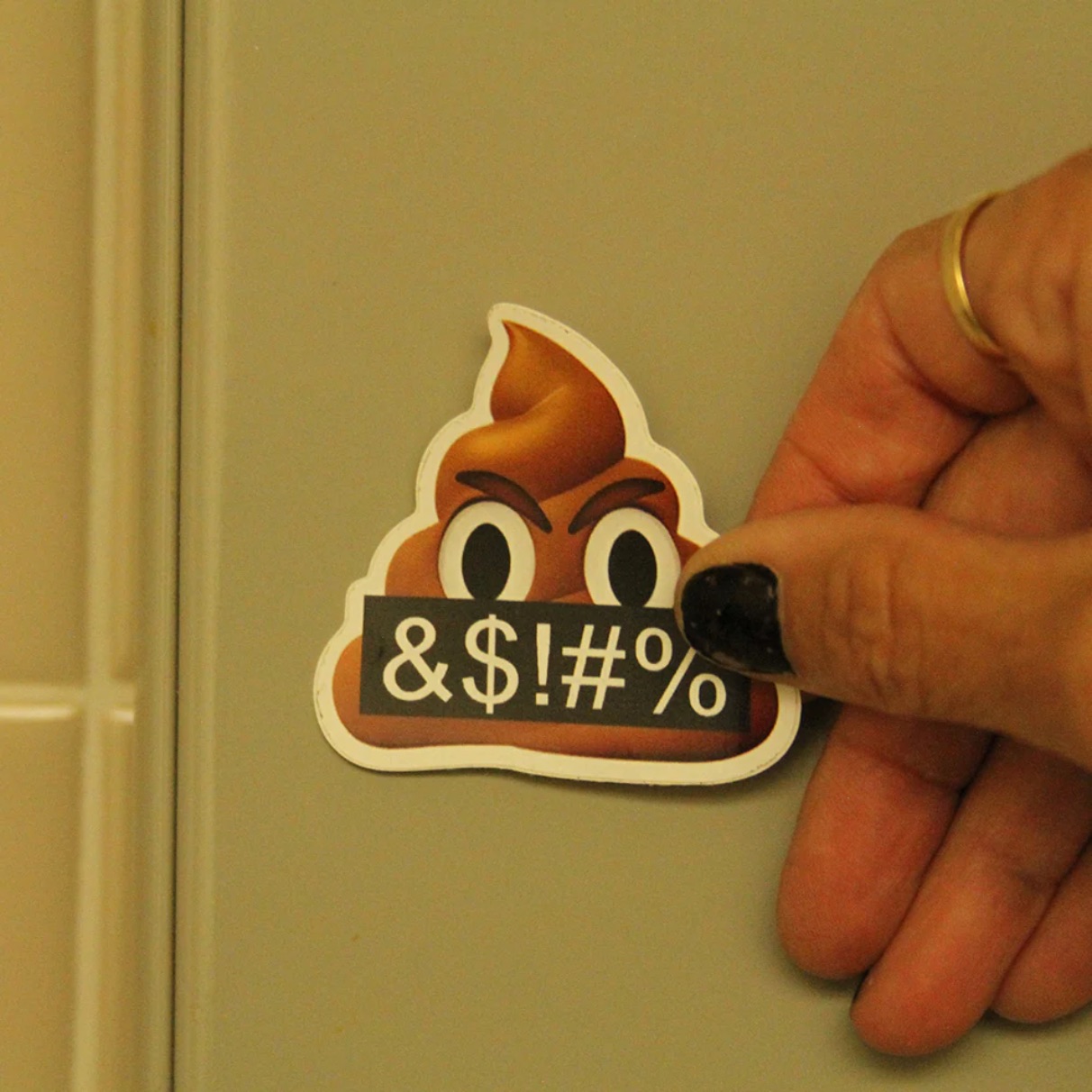 A hand with black nail polish and a gold ring holds an angry poop emoji sticker against a white door. The sticker shows a scowling poop emoji holding a black sign reading "&$!#%".